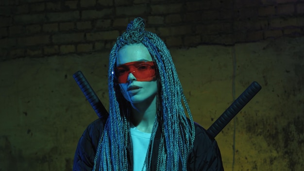 Chica con rastas y katanas en gafas rojas posando contra una pared de ladrillos de neón