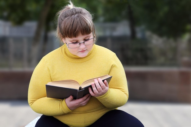 Chica regordeta en anteojos se sienta sola en el parque y libro de lectura reflexivo