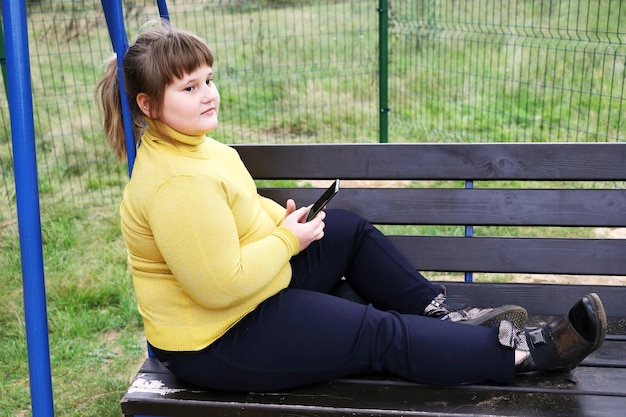 Una chica regordeta y pensativa con suéter amarillo se sienta sola en un banco de columpio al aire libre con un teléfono inteligente