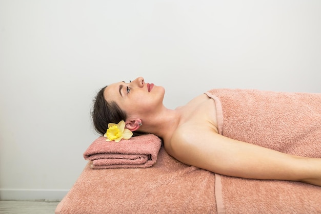 Una chica relajada con una flor en el pelo está acostada boca arriba en la mesa de masajes esperando un masaje Hermosa chica en el spa Concepto de cuidado corporal