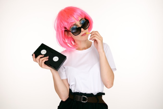 Chica retro de las gafas y peluca rosa con casete negro sobre fondo blanco.