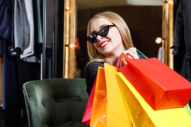 Chica rica y feliz compradora rubia con gafas de sol sosteniendo muchas bolsas de compras coloridas en la tienda de moda Concepto de venta de Black Friday o Cyber Monday