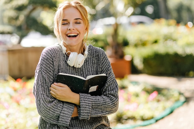 Chica riendo en suéter de punto sosteniendo libro de texto Retrato al aire libre de una adorable estudiante en auriculares escalofriante en un día soleado