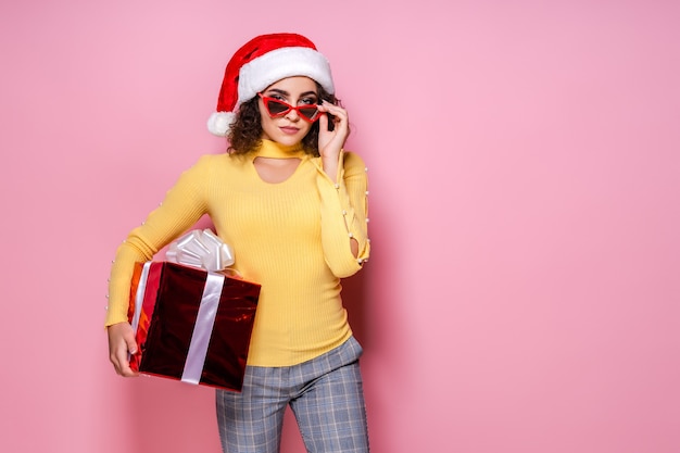 Chica rizada con sombrero de Papá Noel y gafas de sol tiene caja de regalo roja mientras se encuentra sobre fondo rosa año nuevo