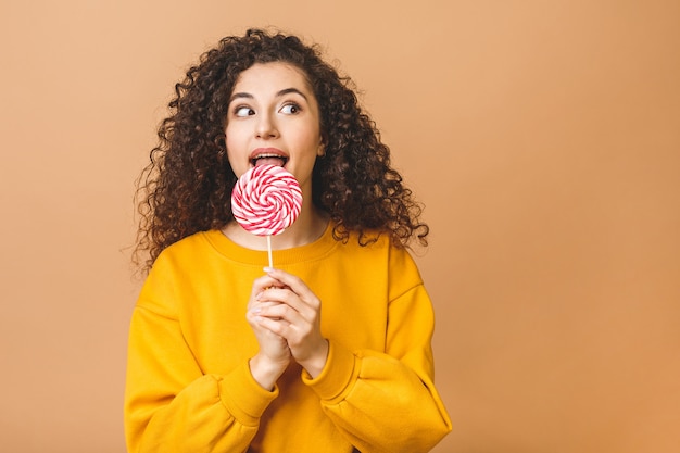 Foto chica rizada sorprendida comiendo piruleta. mujer modelo de belleza con dulce dulce piruleta colorido rosa, aislado sobre fondo beige.