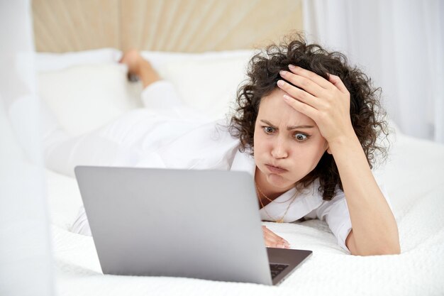 Chica rizada usando una computadora portátil sentada en la cama blanca navegando o charlando con amigos en línea