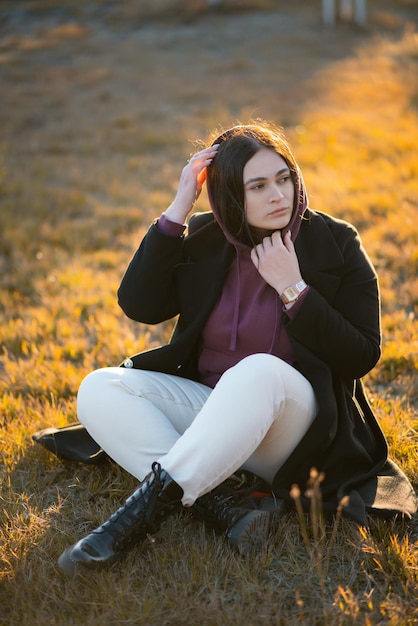 Chica con ropa de abrigo sentada en el césped del parque y posando mirando hacia un lado