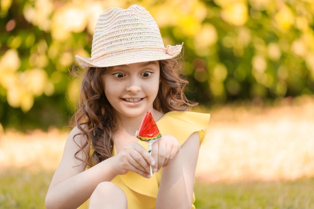 Chica con ropa amarilla y un sombrero se sienta en la hierba en el parque en verano con una paleta en forma de rodaja de sandía. Foto de alta calidad