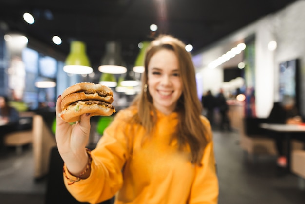 Chica en ropa brillante mantiene una apetitosa hamburguesa grande en el fondo de un restaurante de comida rápida.
