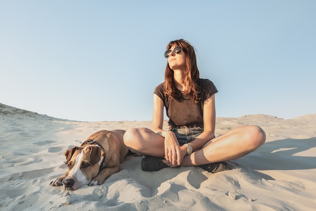 Chica en ropa casual de senderismo y cachorro de Staffordshire Terrier disfrutando de un caluroso día de verano. Hermosa mujer joven con gafas de sol descansa con perro en la playa o el desierto.