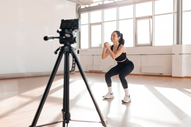 Una chica con ropa deportiva se agacha en el gimnasio y graba el entrenamiento con una cámara de video en un trípode.