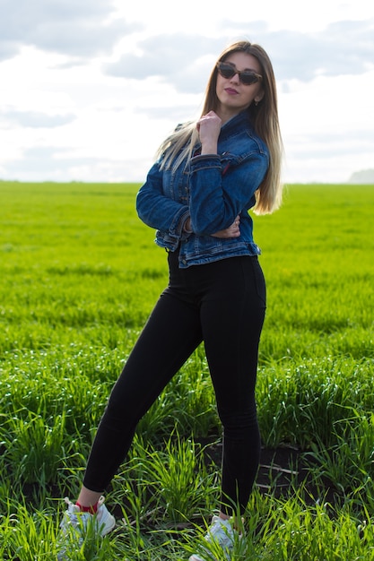 Una chica en ropa de mezclilla con un cabello hermoso se encuentra en un campo verde con una mirada de ensueño en su rostro.