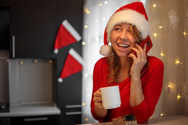 Chica en ropa roja en la cocina hablando por un teléfono inteligente en la víspera de Navidad