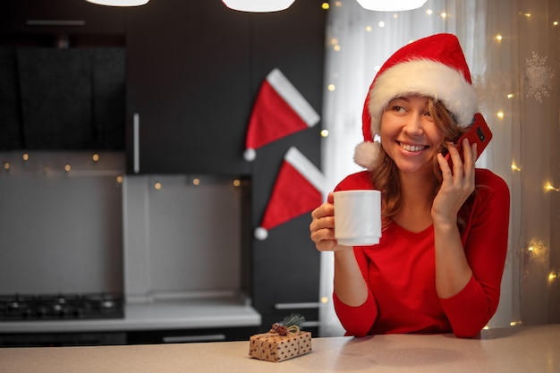 Chica en ropa roja en la cocina hablando por un teléfono inteligente en la víspera de Navidad