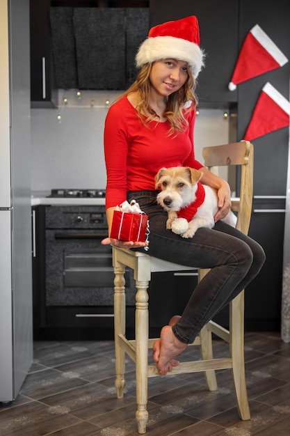 Chica en ropa roja en la cocina muestra un regalo a un perro en la víspera de Navidad