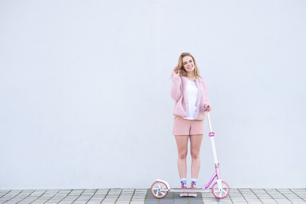 Chica en ropa rosa se encuentra con un scooter en el fondo de una pared blanca, mira a la cámara y sonríe