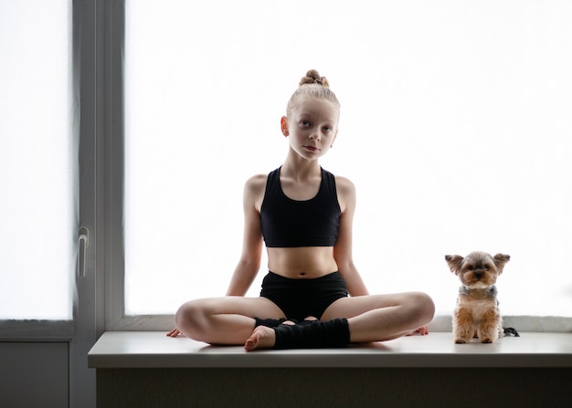 Chica rubia de 8 años haciendo gimnasia en casa sentada en la ventana junto a un cachorro