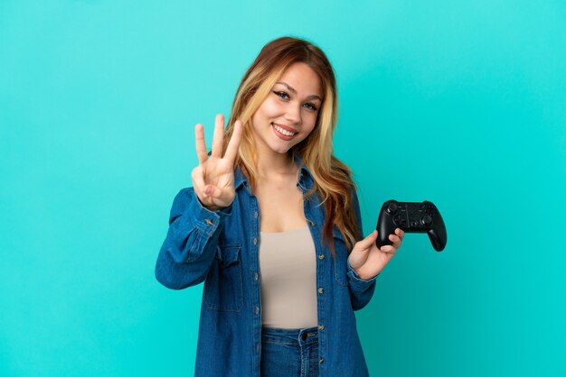 Chica rubia adolescente jugando con un controlador de videojuegos sobre pared aislada feliz y contando tres con los dedos