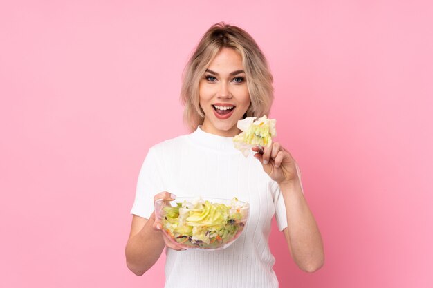 Chica rubia adolescente sosteniendo una ensalada sobre pared rosa