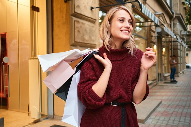Chica rubia alegre en suéter de punto con bolsas de compras en el hombro felizmente mirando hacia otro lado en la calle de la ciudad