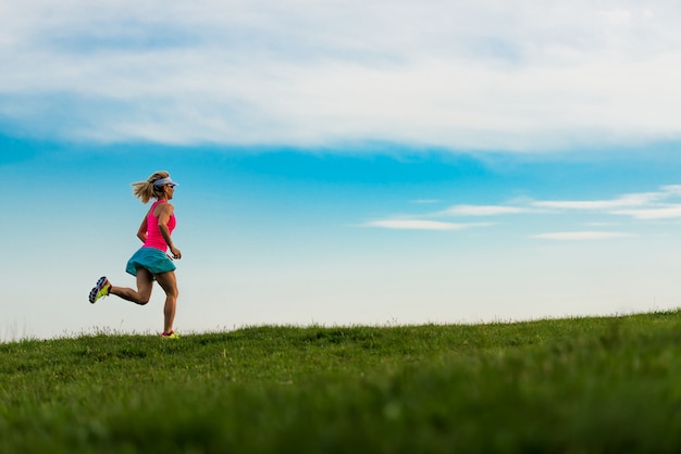 Chica rubia atleta corre en un prado verde en las colinas