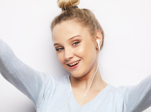 Una chica rubia con auriculares escuchando música tomando fotos hace un autorretrato en un smartphone con ropa informal sobre fondo blanco