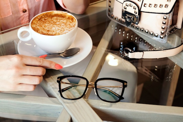 Chica rubia bebe café en la cafetería, gafas, bolso. Blogger aplanada. Café de la mañana en la cafetería.