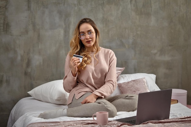 Chica rubia blogger en gafas ropa casual con tarjeta de plástico blanco sentado en la cama con la computadora portátil...