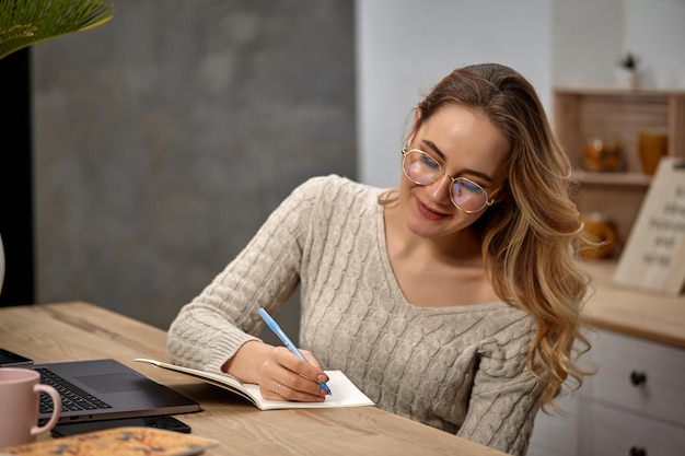 Chica rubia blogger en gafas suéter beige sonriendo sentado en la cocina en la mesa de madera va a escribir...