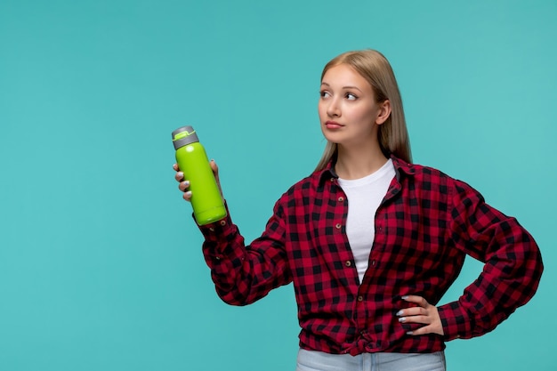 Chica rubia bonita del día internacional de los estudiantes en camisa roja a cuadros con matraz verde