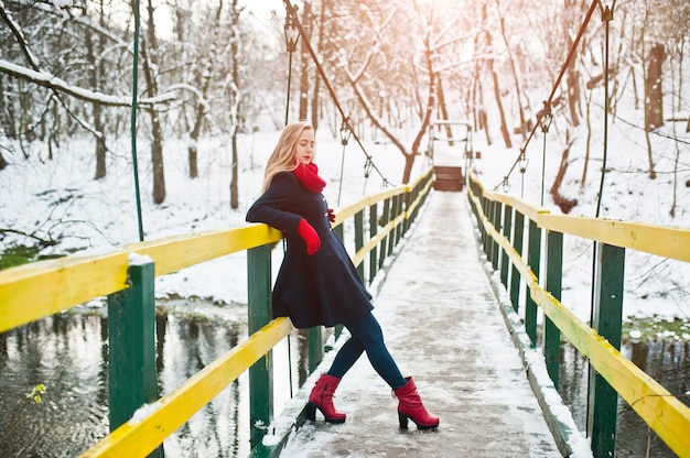 Chica rubia con bufanda roja y abrigo caminando sobre el puente en el parque el día de invierno.