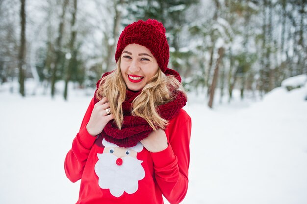 Chica rubia en bufanda roja, sombrero y suéter santas posando en el parque en día de invierno.