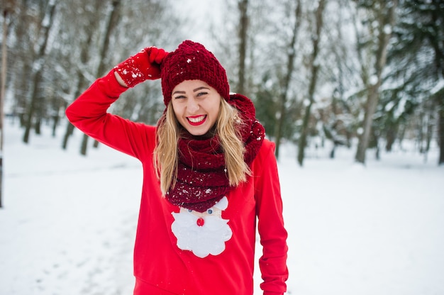 Chica rubia en bufanda roja, sombrero y suéter santas posando en el parque en día de invierno.