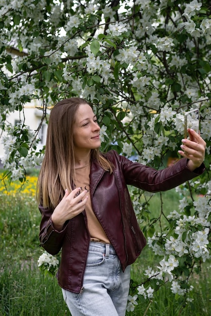 Una chica rubia caminando en el parque hace un selfie en un teléfono inteligente y se ríe de que la temporada de primavera es el florecimiento del manzano, la gente usa la tecnología