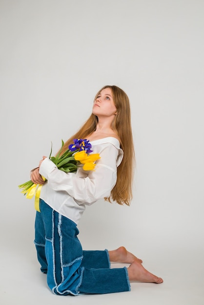 Una chica rubia con una camisa blanca y jeans azules sostiene un ramo de tulipanes e iris sentado