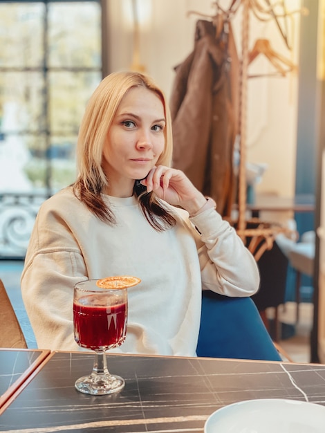 Una chica rubia caucásica alegre con un suéter ligero se sienta en un restaurante cerca de la ventana en una mesa