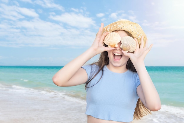 Chica rubia cubriendo sus ojos con dos conchas y sonriendo con risa en una playa de aguas turquesas