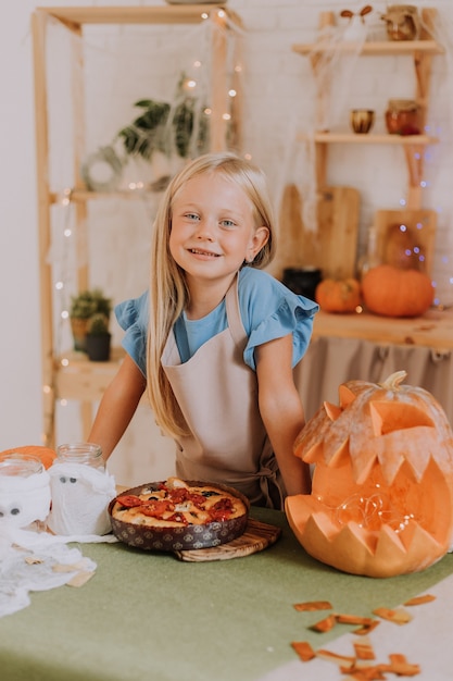 Chica rubia en delantal en la cocina decorada con calabazas para Halloween prepara un pastel de focaccia