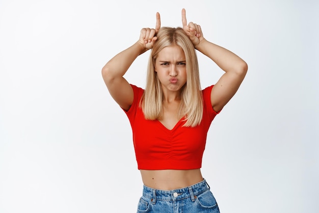 La chica rubia descarada muestra un gesto de cuerno de toro siendo obstinado haciendo pucheros y mirando seriamente a la cámara fondo blanco Copiar espacio