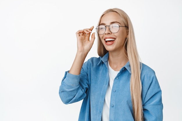 Foto chica rubia despreocupada riendo, sonriendo y mirando feliz, ponerse gafas, de pie en traje casual en blanco.