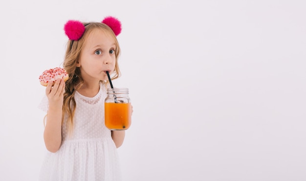 Chica rubia divertida comiendo un donut y bebiendo jugo de fruta en un fondo blanco