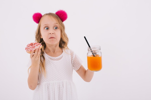 Chica rubia divertida comiendo un donut y bebiendo jugo de fruta en un fondo blanco