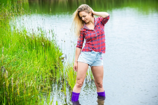La chica rubia se divierte en el río con botas de goma en el campo