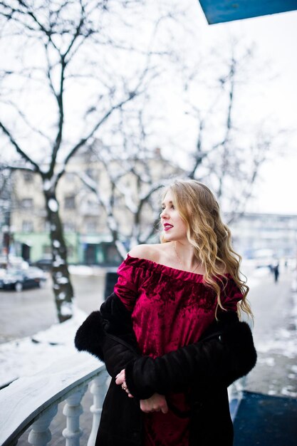 Chica rubia de elegancia en vestido de noche rojo y abrigo de piel en las calles de la ciudad en el día de invierno