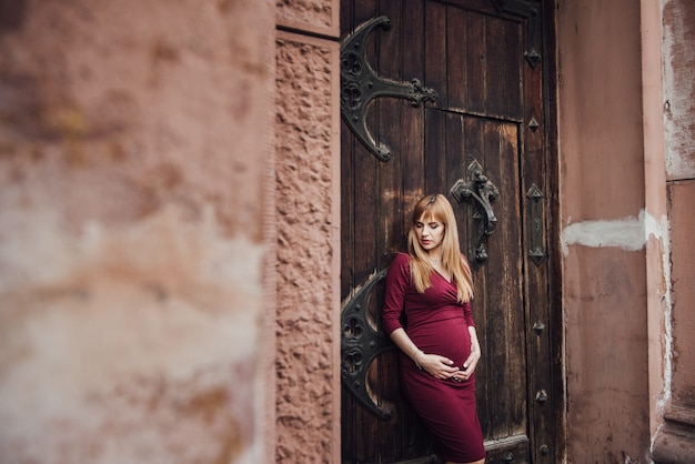 Chica rubia embarazada en vestido burdeos. En la gran ciudad Pelo largo. 9 meses de espera. La felicidad de ser madre. Contra el telón de fondo de un hermoso edificio antiguo.