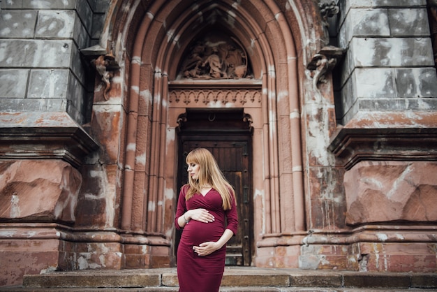 Chica rubia embarazada en vestido burdeos. En la gran ciudad Pelo largo. 9 meses de espera. La felicidad de ser madre. Contra el telón de fondo de un hermoso edificio antiguo.
