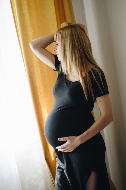 Chica rubia embarazada en un vestido negro en casa. Esperando un milagro. El embarazo. Amor y esperanza. Habitación luminosa.