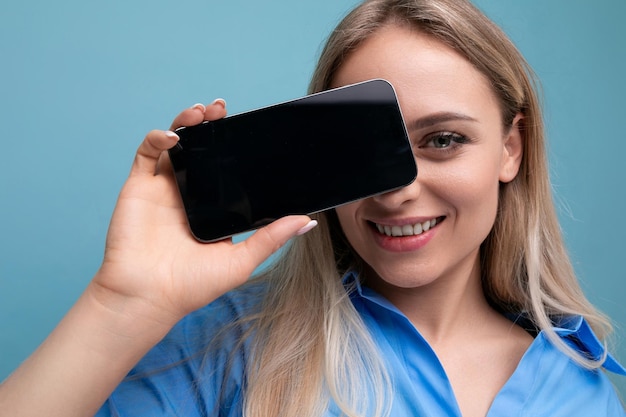 Chica rubia con estilo sonriente sosteniendo la pantalla del teléfono inteligente con maqueta para la página web cerca de su cara en azul