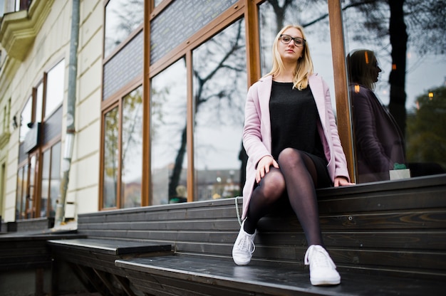 Chica rubia en gafas y abrigo rosa, túnica negra sentada en el banco en la calle.