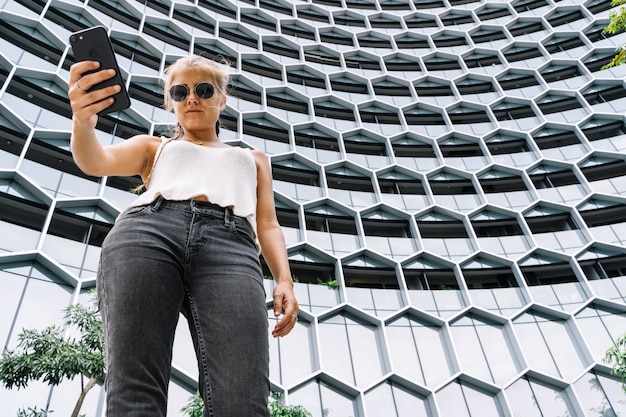 Chica rubia con gafas de sol de pie delante de un edificio moderno usando un móvil